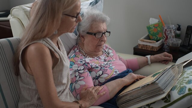 In a senior living facility, a daughter and her elderly mother smile and laugh as they reminisce while flipping through a photo album filled with memories.