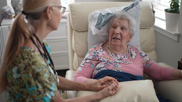 A nurse holds the hand of an elderly woman while engaging in a friendly conversation in a senior living facility, creating a warm atmosphere of support.