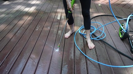 A mature woman uses a pressure washer to clean a wooden deck, removing dirt and stains for better outdoor living space. The sun shines as she works.