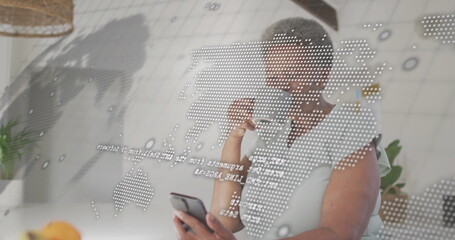 Holding smartphone and coffee mug, woman analyzing world map projection in home kitchen with plants