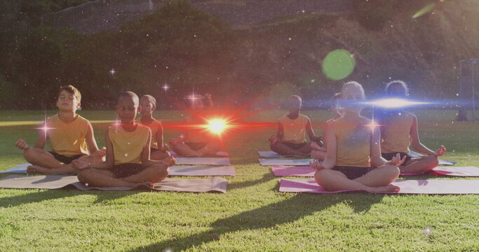 Meditating children sitting cross-legged on yoga mats in park on grassy lawn, wearing yellow tops