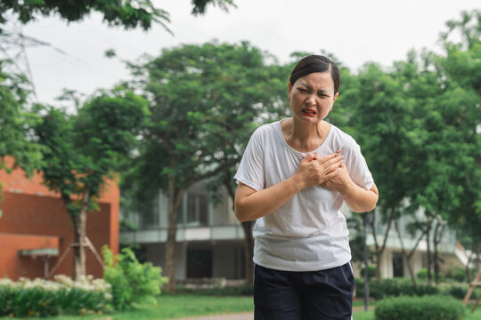 Woman clutches chest experiencing heart pain while outdoors in a green park setting looking distressed