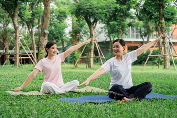 Two women practice yoga outdoors sitting cross-legged with arms outstretched in a serene park setting