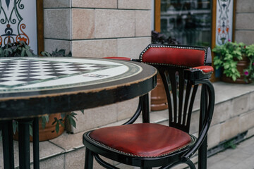 cozy outdoor cafe features chess table at forefront with an empty red chair, inviting patrons to enjoy leisurely game. lush greenery adds vibrancy to atmosphere. close up.