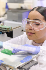 Young female researcher in a university lab performs 3D printing tests. She wears a white lab coat, nitrile gloves, and safety glasses while carefully operating the printer