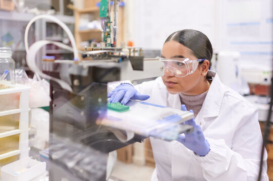 Young Latina female scientist works with lab coat and safety glasses at 3D printer in laboratory