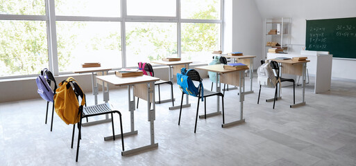 Interior of empty classroom with desks, backpacks on chairs and blackboard