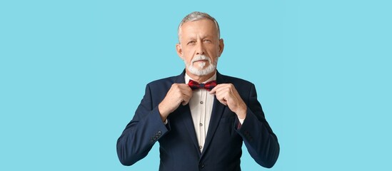 Handsome mature man in stylish formal suit fixing bow tie on blue background