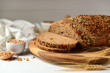 Wooden board with sliced loaf of bread, wheat spikelets and grains on white table