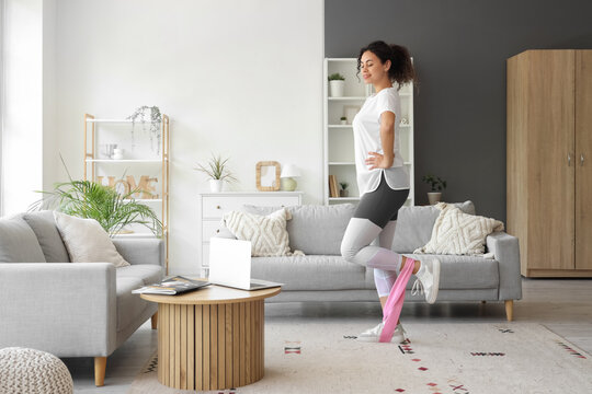 Sporty African-American woman with laptop and resistance band exercising at home
