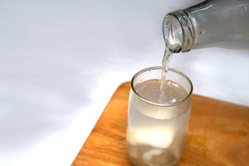 Pouring Coconut water in a glass with straw on white wooden table over white background. Fresh detox coconut juice. Selective focus.