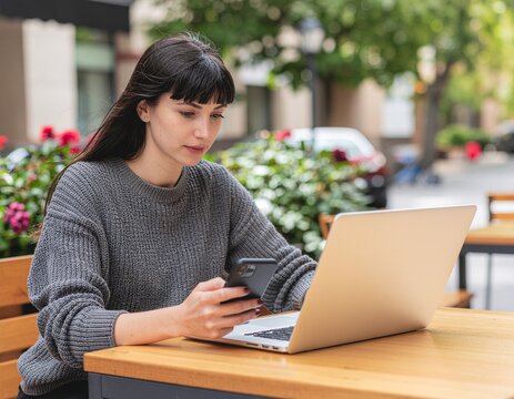 A young woman working remotely on her laptop and smartphone at an outdoor city cafe, representing flexible work and modern lifestyle. - Powered by Adobe
