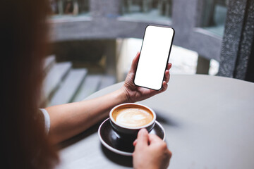 Mockup image of a woman holding and using mobile phone with blank desktop screen while drinking coffee in cafe