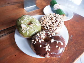 Three donuts served in white plate with various flavor. peanut, chocolate and matcha, side view with brown background
