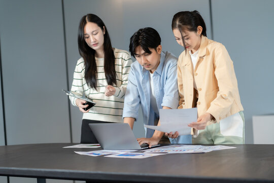 Group of asian coworkers standing around a table during a team brainstorming session, reviewing ideas and charts together to support project success through collaborative team brainstorming
