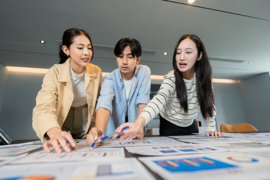 Three asian coworkers discussing ideas and pointing at data charts during a business meeting, actively participating in a collaborative decision process to ensure a successful collaborative decision