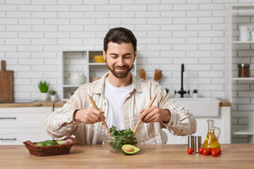 Handsome man preparing tasty kale salad with avocado and tomatoes at wooden table in kitchen