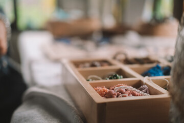 Close up shot of a jewelry designer's desk in a house with large windows and lush greenery outside. Beads, tools, and jewelry are on the desk. Work process, small business.
