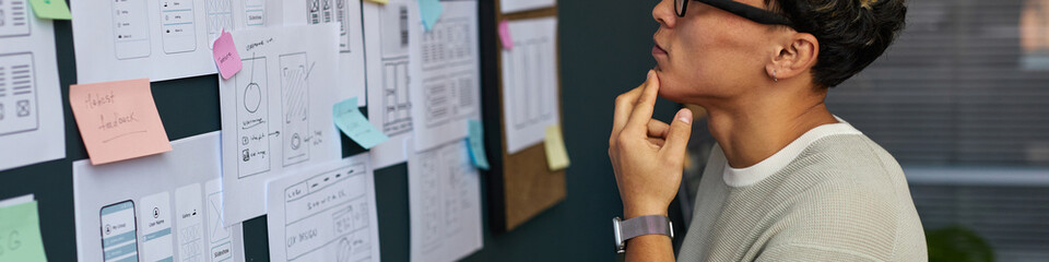 Fototapeta premium Young adult Asian woman analyzing wireframe sketches and sticky notes on wall, standing in modern office, wearing glasses, planning user interface design for digital project