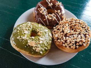 3 Donuts with varios flavour, chocolate, peanut and green tea donuts on the table, green background, top view, served by white plate
