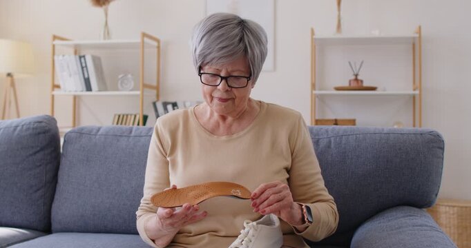 Old woman suffering from flat feet, bought orthopedic shoes by doctors recommendation, touching new orthotic insole, choosing comfortbale footwear to wear, sitting on comfortbale sofa