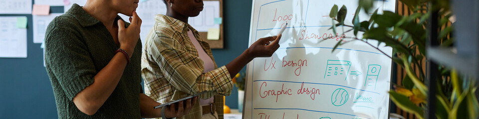 Young adult Asian man holding tablet, standing beside young adult Black woman pointing at...