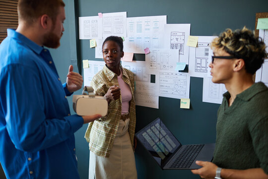 Multiethnic group of young adults collaborating on project, Black woman gesturing while discussing ideas with Caucasian man holding virtual reality headset and Asian man using laptop