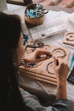 Back view of a young brunette woman, a jewelry designer. She is sitting at her workspace, where she has tools and various beads that she uses to create jewelry. Home based small business, startup.