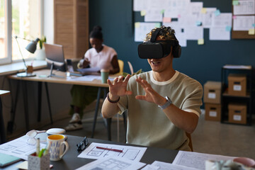 Young adult Asian man wearing virtual reality headset and brainwave sensors, interacting with digital interface at desk while Black woman working in background office setting