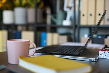 Fototapeta premium Closeup showing laptop, pink mug, and yellow notebook on desk in modern office workspace, blurred shelves with folders and plants in background, no people visible