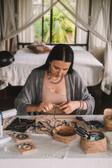 Portrait of a young brunette woman jewelry designer. She is sitting at a work table on which lie tools and various beads from which she creates jewelry. Home small business, startup. Creative hobby.