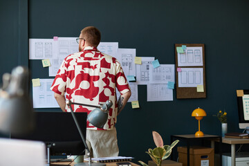 Caucasian young adult man standing in modern office analyzing mobile app wireframes and sketches pinned on wall, working on user interface design project surrounded by digital devices