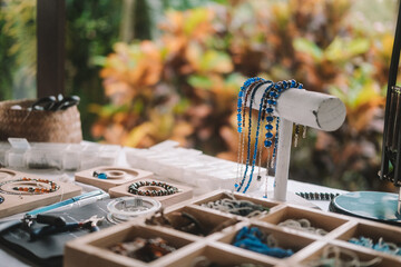 Close up shot of a jewelry designer's desk in a house with large windows and lush greenery outside. Beads, tools, and jewelry are on the desk. Work process, small business.