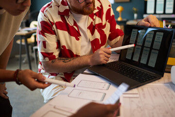Young adult Caucasian man and young adult Asian woman collaborating on user interface design, discussing wireframes displayed on laptop screen, holding markers and reviewing sketches