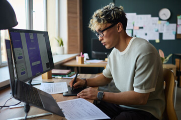 Young adult Asian man working at desk using digital pen and tablet designing mobile app interface on computer monitor, surrounded by sketches and documents in modern office