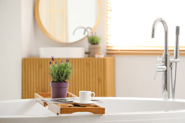 Stand with pot of lavender flowers, magazine and cup of coffee on bathtub in modern bathroom, closeup