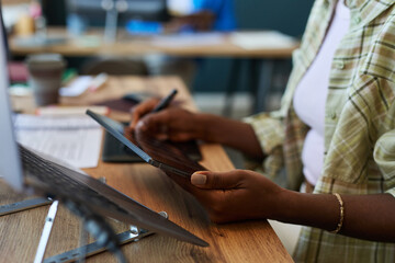 Young adult Black woman using digital tablet with stylus at desk, focusing on creative work in modern office setting, hands and partial upper body visible, technology in use