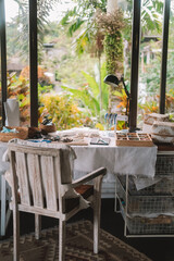 Medium shot of a jewelry designer's desk in a house with large windows and lush greenery outside. Beads, tools, and jewelry are on the desk. Work process, small business.