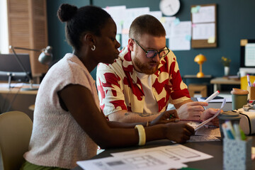 Black woman and Caucasian man collaborating at desk, reviewing documents and discussing project...