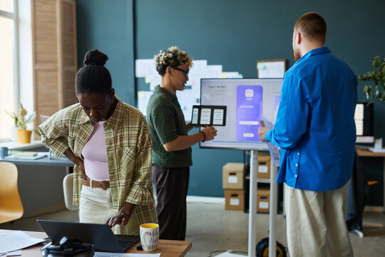 Young Black woman working on laptop while multiethnic group of young adults collaborating on digital project near interactive display in modern office workspace - Powered by Adobe