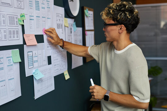 Young adult Caucasian man standing at wall, organizing paper wireframes and sticky notes, holding marker, developing user interface design, focusing on creative planning process