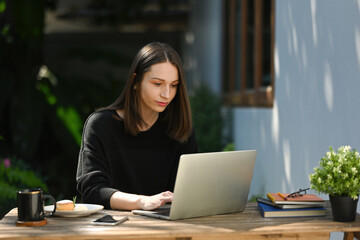 Fototapeta premium Concentrated young Caucasian woman working on a laptop at an outdoor wooden table, enjoying a coffee and a snack, representing remote work or freelance lifestyle