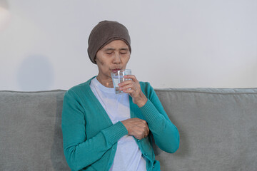 An elderly woman with cancer sits on a sofa at home, wearing a headwrap and drinking water. She...