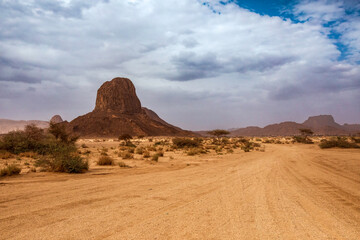 A wide, low-angle shot captures a vast desert plain leading towards a striking, isolated rock formation that dominates the horizon. The sandy foreground shows tracks, suggesting passage. Sparse vegeta