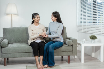 An Asian daughter gently holds her elderly mother’s hand as they sit on a sofa at home, sharing a tender moment of comfort, care, and emotional connection in the living room