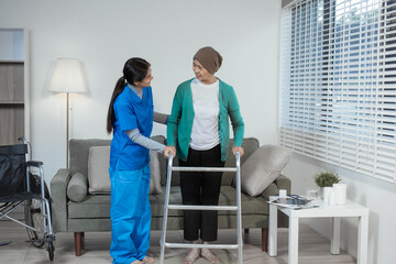 A young Asian nurse assists an elderly female cancer patient, wearing a headscarf, with using a...
