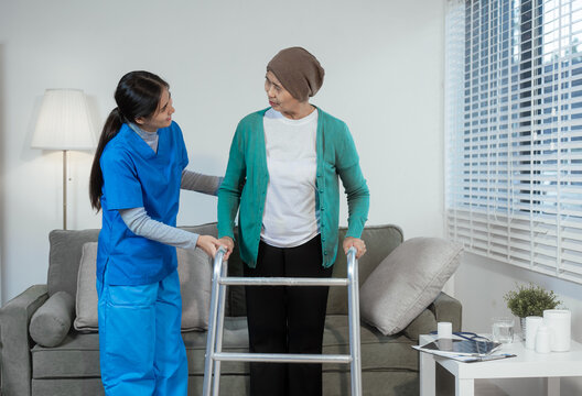 A young Asian nurse assists an elderly female cancer patient, wearing a headscarf, with using a walker. The patient sits on a sofa, preparing for rehabilitation and mobility training in a caring - Powered by Adobe