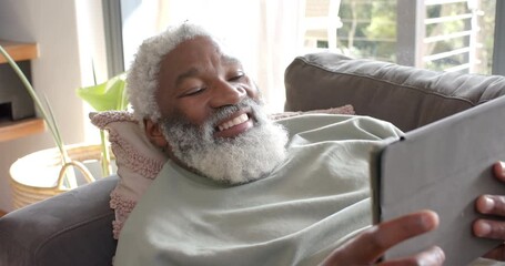 Upon reclining on sofa, African American senior man tapping tablet and speaking for reading at home - Powered by Adobe