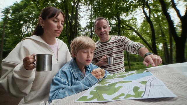 Family of three gathered by car hook having tea with snacks and looking at paper map while young man pointing at it and explaining further route of their trip