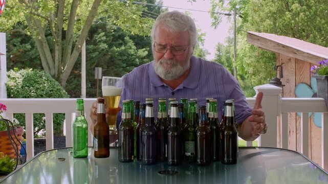 An elderly man sits at a table in his backyard, sampling different beers. He examines each bottle and enjoys the process of tasting, surrounded by various beer bottles.